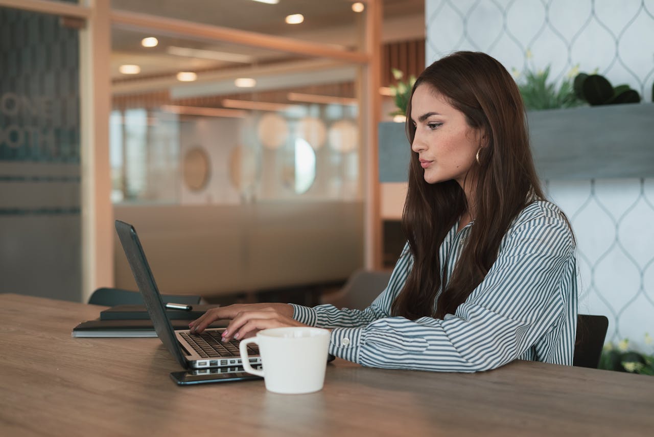 A professional woman working on her laptop at a modern office with a coffee cup nearby.