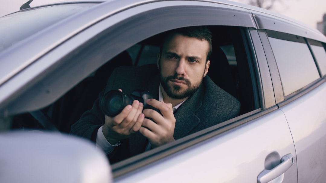 Young private detective man sitting inside car and photographing with slr camera